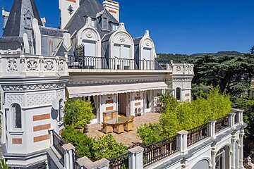 An ornate, multi-story villa with decorative balconies and a large outdoor patio featuring a dining set and lush plants, under a clear blue sky.