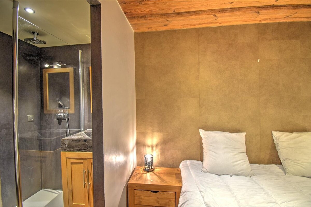 Cozy bedroom with bed, white pillows, and lamp next to a modern glass-enclosed bathroom with shower and sink. Earthy tones, wooden beams.