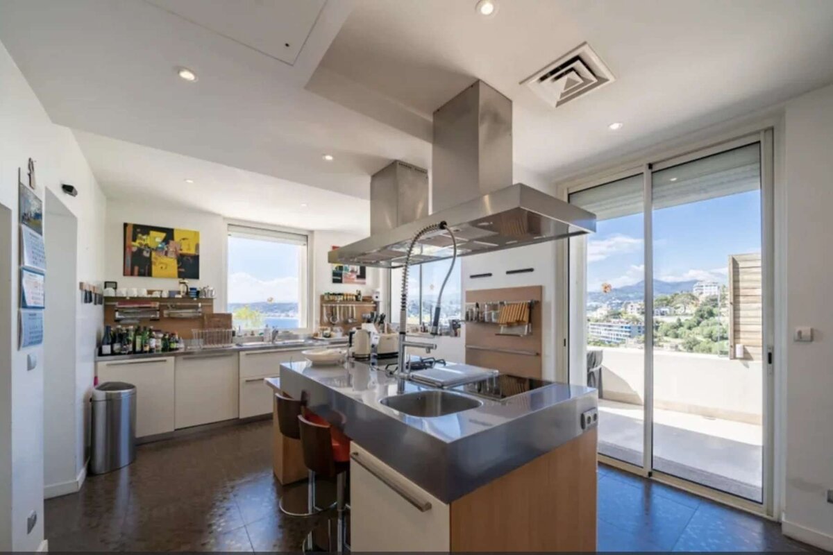A modern kitchen features a large stainless steel island, sleek appliances, and expansive windows/sliding doors offering a panoramic city and sea view.