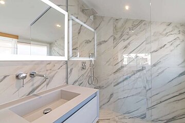A modern bathroom with white marble walls and floor, featuring a built-in sink, lighted mirror, and a glass-enclosed shower with a rainfall showerhead.