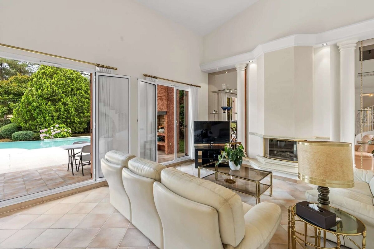 Bright living room with cream sofas, a fireplace, and large sliding glass doors opening to a lush garden with a swimming pool.