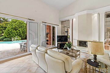 Bright living room with cream sofas, a fireplace, and large sliding glass doors opening to a lush garden with a swimming pool.