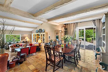 A rustic open-plan living and dining room with terracotta floors, exposed beams, traditional furniture, and French doors opening to a lush garden.