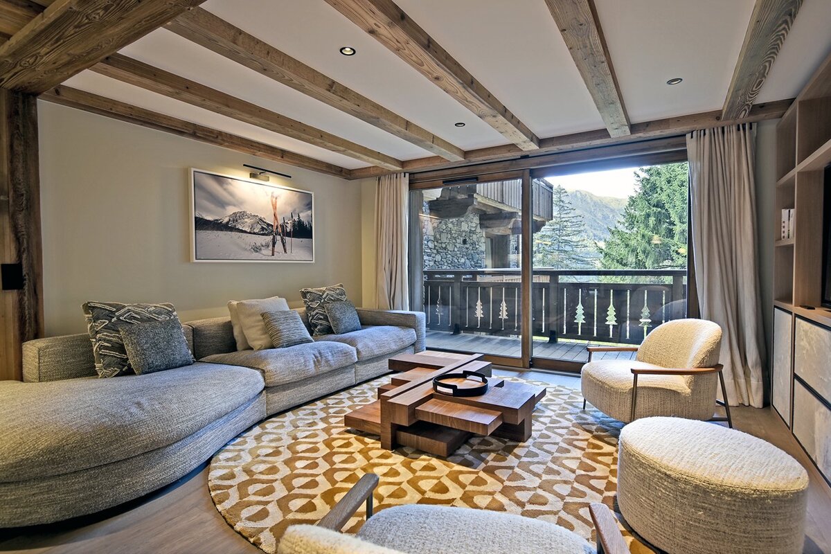 Cozy chalet living room with a curved grey sofa, wooden ceiling beams, a patterned rug, and glass doors opening to a balcony.