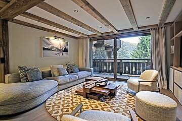 Cozy chalet living room with a curved grey sofa, wooden ceiling beams, a patterned rug, and glass doors opening to a balcony.