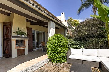 Sunny house patio with a built-in BBQ, outdoor sectional sofa, and lush green plants under a bright blue sky.
