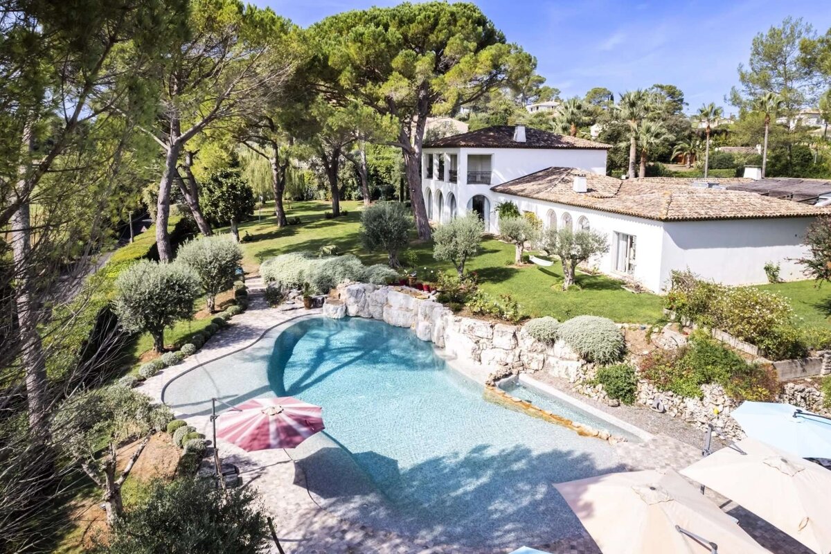 An aerial view of a beautiful white Mediterranean villa with a freeform swimming pool, nestled in a lush green garden with tall pine trees.