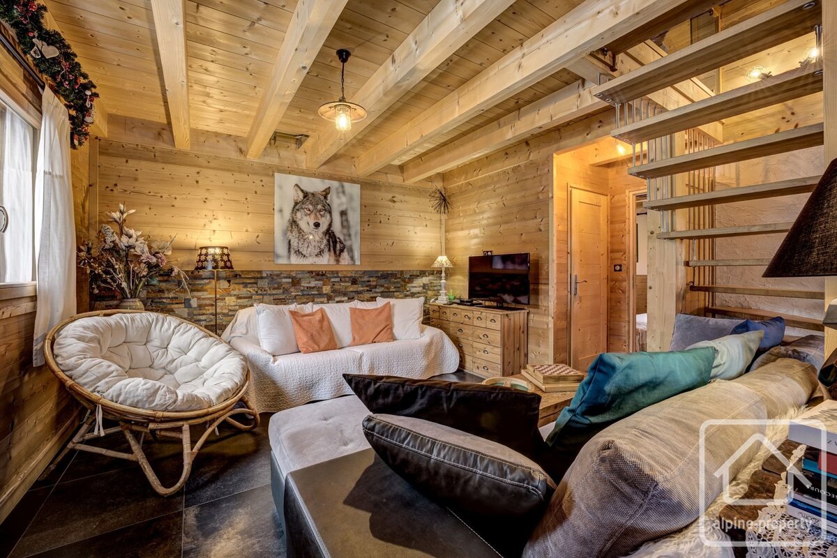 A cozy chalet-style living room with wood paneling, exposed beams, a papasan chair, and a sofa under a wolf painting. A wooden staircase is visible.