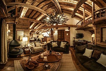 A warm, luxurious chalet living room with a vaulted wood ceiling, exposed beams, fireplace, comfy sofas, and a rustic aesthetic.