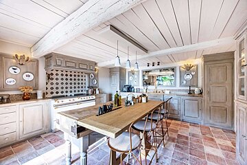 A spacious rustic kitchen boasts a long wooden island with seating, distressed gray cabinets, a tiled backsplash, and a white beamed ceiling over a tiled floor.