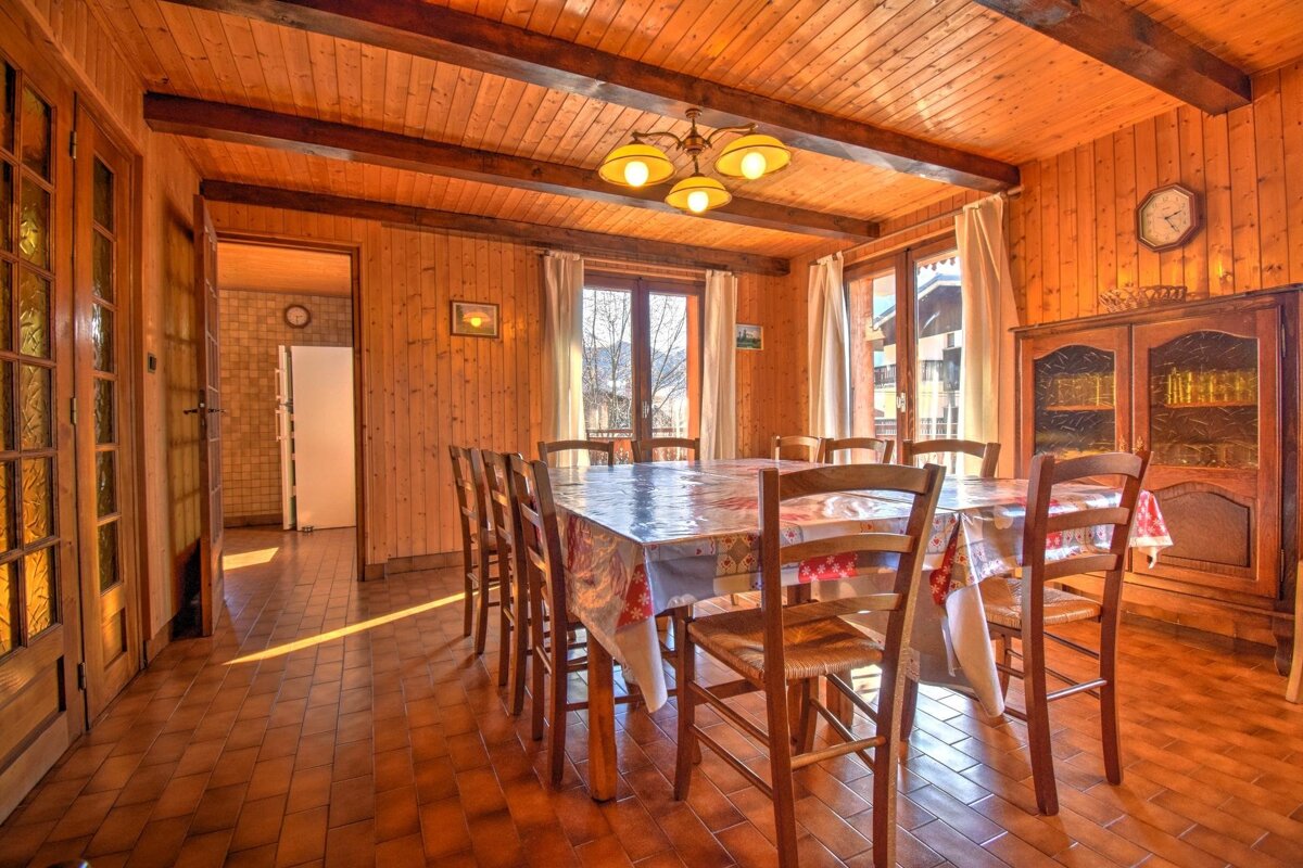 Rustic dining room with wood paneling on walls and ceiling, a long table, tiled floor, and natural light from windows.