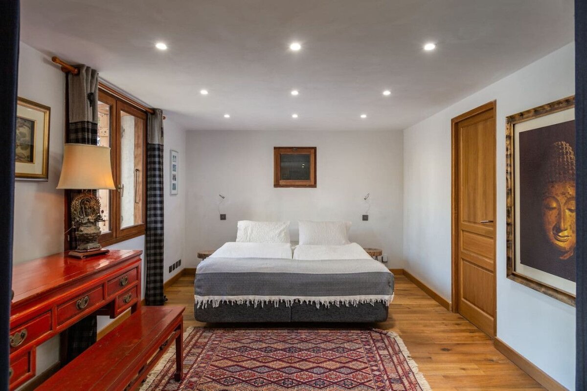 A cozy bedroom with a large bed, wooden floors, an oriental rug, red dresser, and window with plaid curtains. Buddha art adds a calm touch.