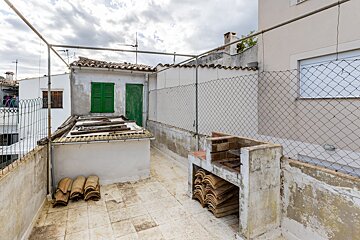 A worn rooftop terrace features a built-in barbecue, stacked tiles, and a building with a green door/shutters, all under a cloudy sky.