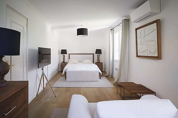 Bright, minimalist white bedroom with a large bed, wooden floors, a TV on a tripod, and a white sofa, creating a clean and modern look.