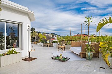 A bright rooftop terrace with wicker furniture, many plants, and a distant city view beneath a partly cloudy sky.