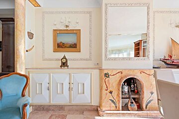 An ornate, classical room with light walls, featuring a landscape painting, antique clock, large mirror, and a decorative fireplace bar next to a blue armchair.