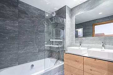 A modern bathroom with grey tiled walls, a bathtub with a glass shower screen, and a double vanity featuring white vessel sinks on a light wood cabinet.
