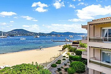A beautiful sunny day at a scenic coastline with a sandy beach, clear blue ocean, many yachts, distant mountains, and modern buildings by the shore.