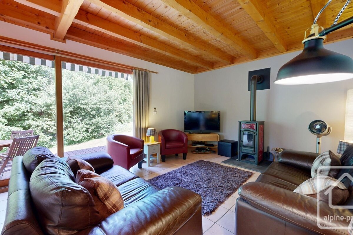 Warm living room with wood-beamed ceiling, brown leather sofas, a wood-burning stove, and a large sliding door opening to green trees.