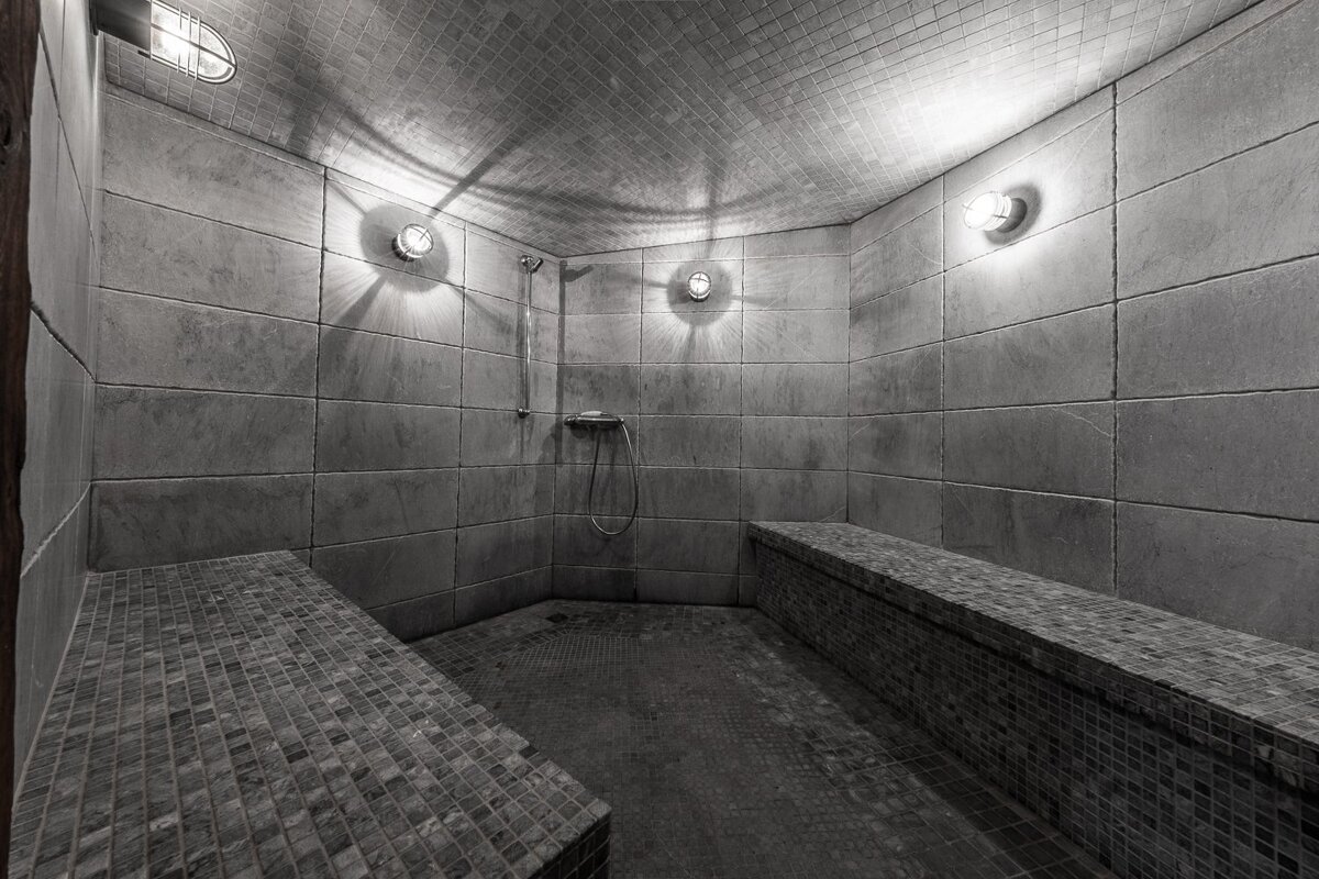 A minimalist grey-tiled steam room featuring mosaic benches, dramatic lighting, and a corner shower.