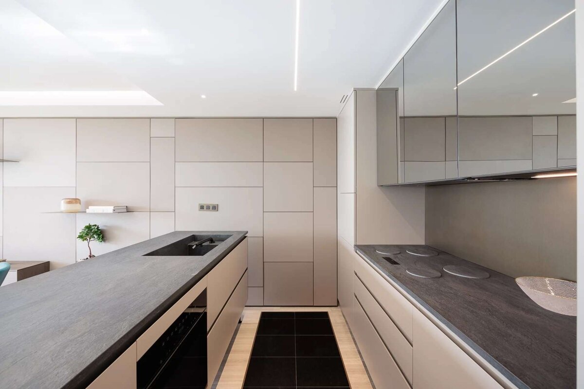A sleek, minimalist galley kitchen features light beige cabinetry, dark grey countertops, and bright linear lighting, creating a clean, modern aesthetic.