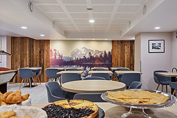 A bright dining room with blue chairs, wooden tables, and a large mountain mural. Croissants and pastries are visible in the foreground.