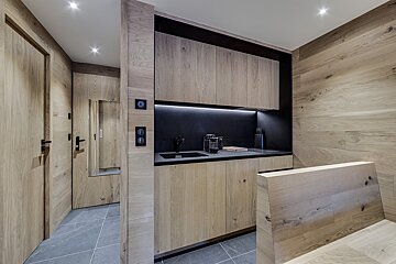 A modern, compact kitchen/bar area featuring light wood cabinetry, dark countertops, and backsplash lighting. A grey tiled floor and matching wood doors are visible.