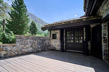 Rustic stone cabin with a wooden deck patio, overlooking pine trees and mountain peaks under a blue sky.