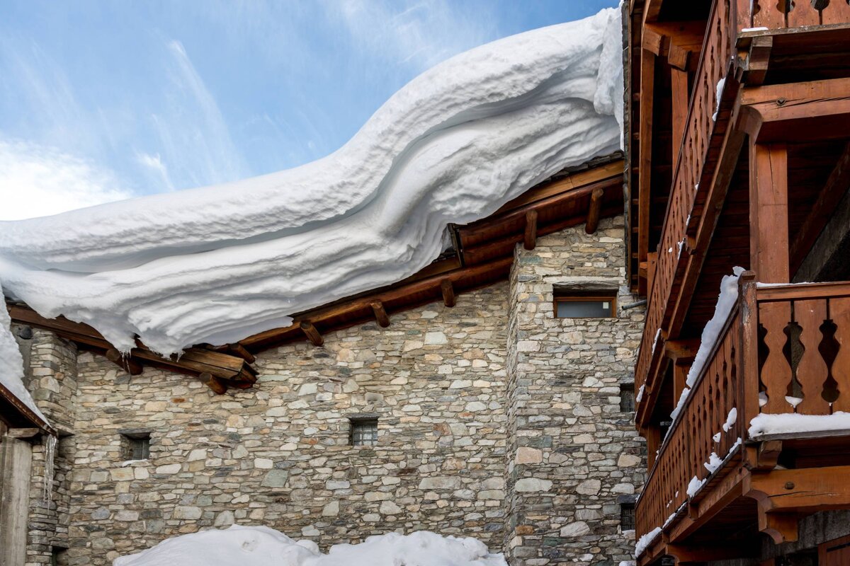 A stone house with a massive, wavy snowdrift overhanging its roof, next to a wooden balcony, under a clear blue sky.