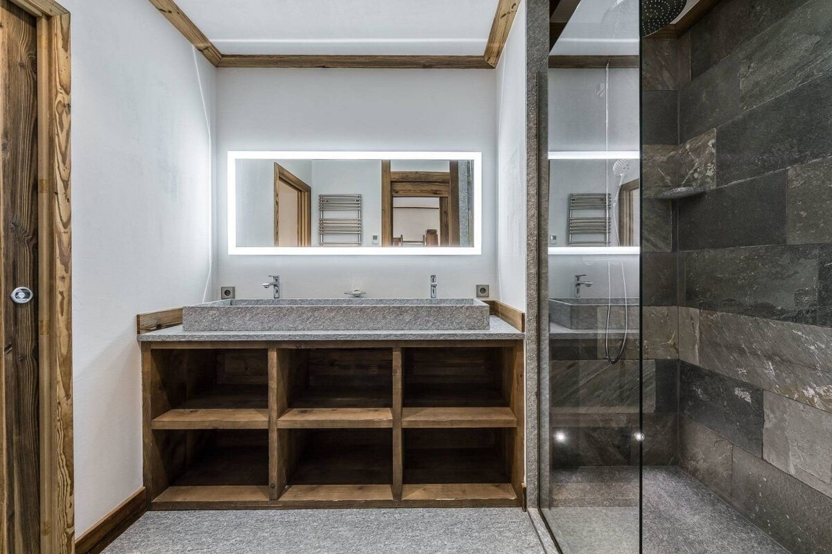A modern rustic bathroom features a double stone sink vanity with a lighted mirror, alongside a dark slate-tiled walk-in shower. Natural wood accents complete the look.