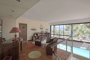 A bright, nautical-themed room with large windows overlooking the sea, palm trees, and a pool below. Features wooden floors, chairs, and ship models.