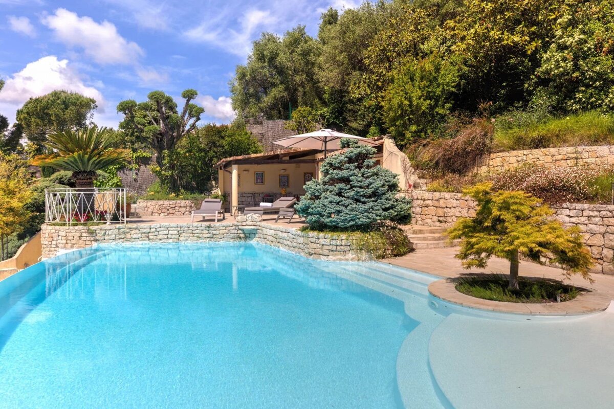 A luxury outdoor swimming pool surrounded by stone patios, lush greenery, and a covered lounge area.