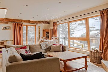 A bright, cozy living room with a sofa and dining area, featuring large windows with a snowy mountain view. Warm wooden accents create a rustic chalet feel.