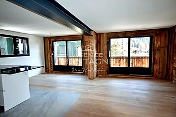 An empty chalet interior with light wood floors, rustic wood-paneled walls, large sliding glass doors, and a kitchen counter.