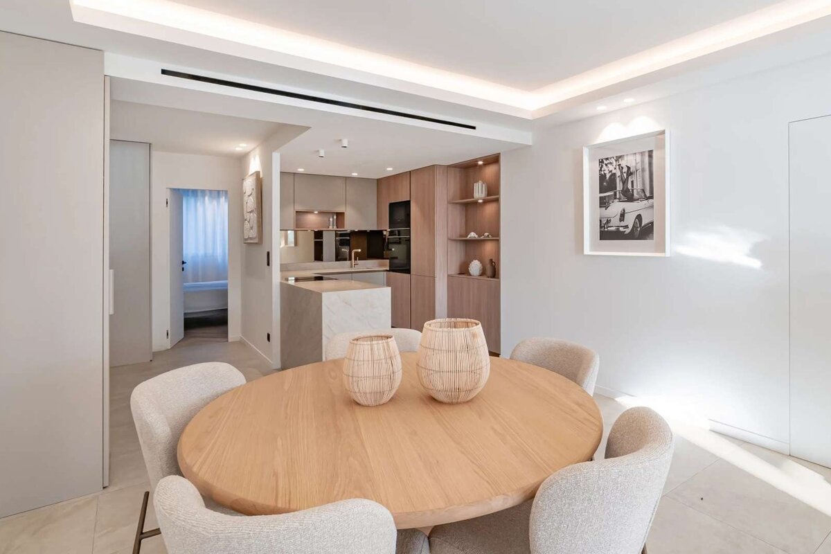 Bright, modern open-plan dining and kitchen area with a light wood table, neutral chairs, and sleek wooden cabinets.
