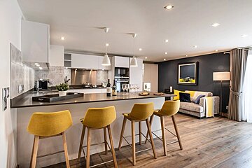 Modern open-plan kitchen and living area featuring a large island with yellow bar stools, sleek white cabinetry, and a cozy lounge with a dark accent wall.