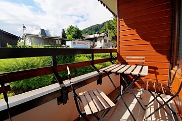 A cozy balcony with a wooden table and chairs, overlooking a European village with green hills and mountains under a partly cloudy sky.