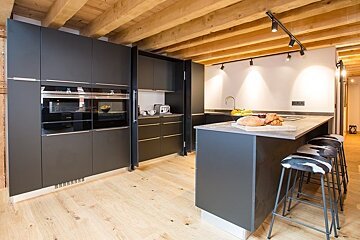 Modern kitchen with dark grey cabinets, rustic wooden ceiling beams, light wood floors, and an island with cowhide barstools.