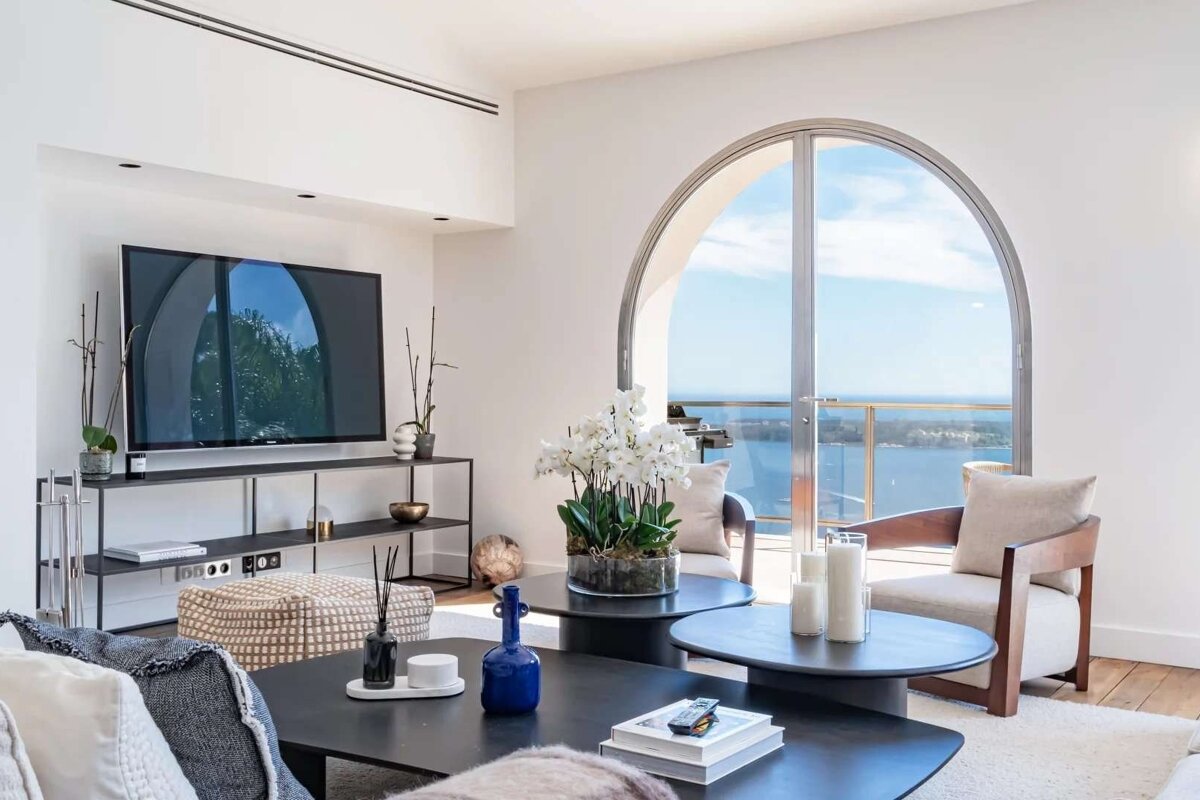 A bright, modern living room with a stunning arched window offering expansive water views. It features a large TV, stylish furniture, and white orchids.