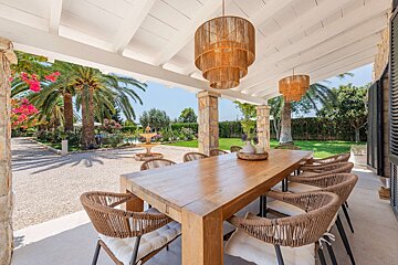 A beautiful outdoor dining area with a long wooden table, woven chairs, and stylish chandeliers, overlooking a lush garden with palm trees and a fountain.