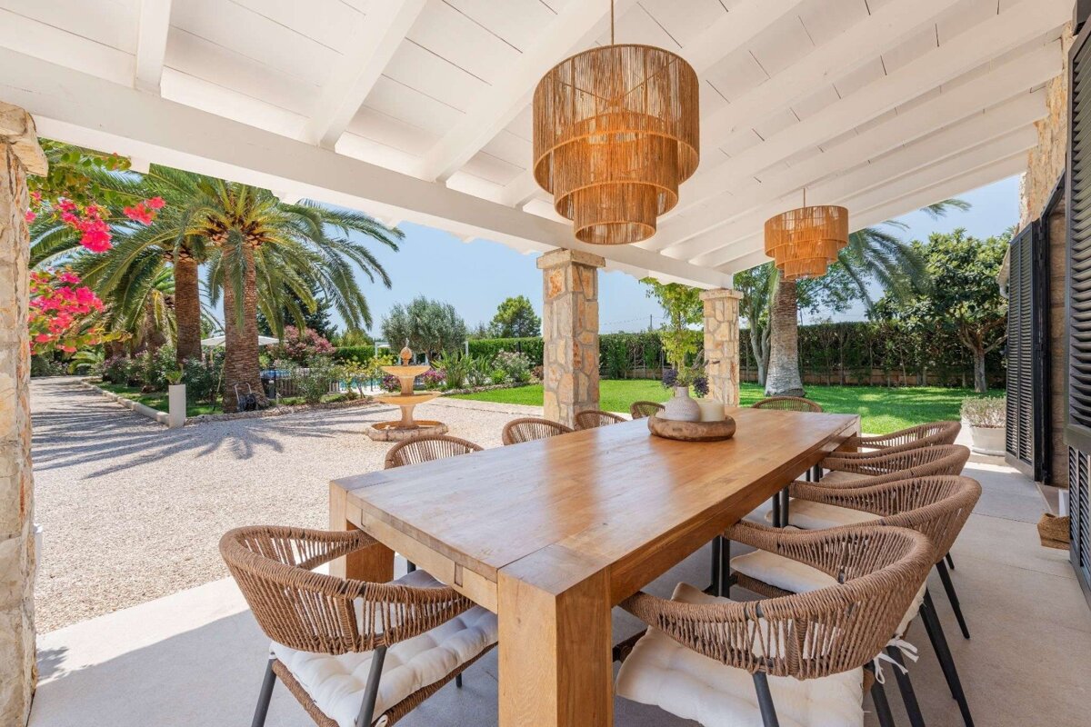 Stylish outdoor dining area with a long wooden table, woven chairs, and rattan chandeliers under a pergola, overlooking a lush garden with palm trees and a fountain.