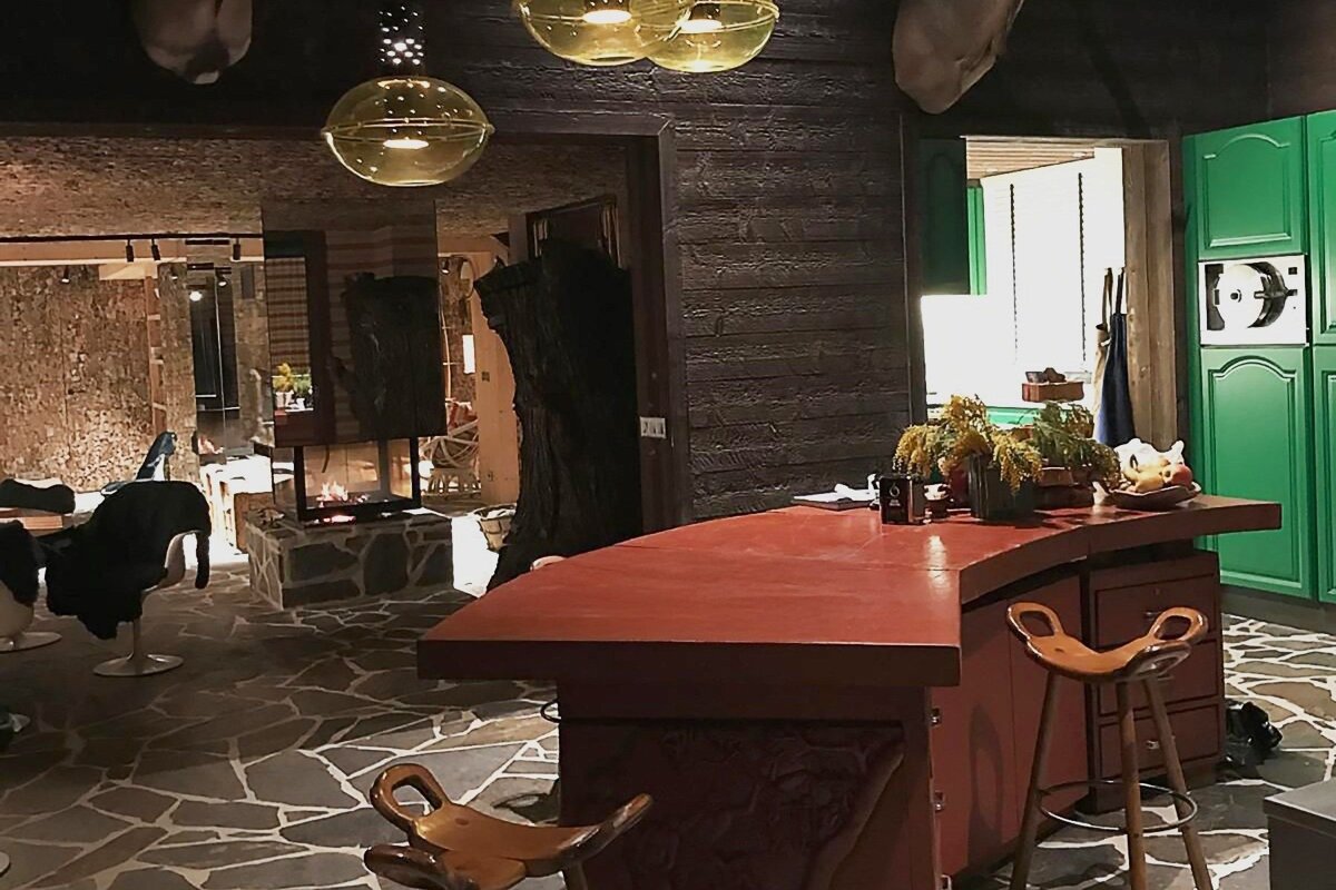 An eclectic interior features a red bar with wooden stools, green kitchen cabinets, a stone floor, and a fireplace, all under gold orb lights.