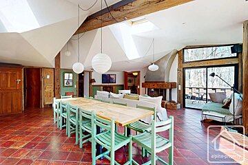 Rustic open-plan interior with a long wooden dining table, green chairs, red tile flooring, and vaulted ceilings with skylights.