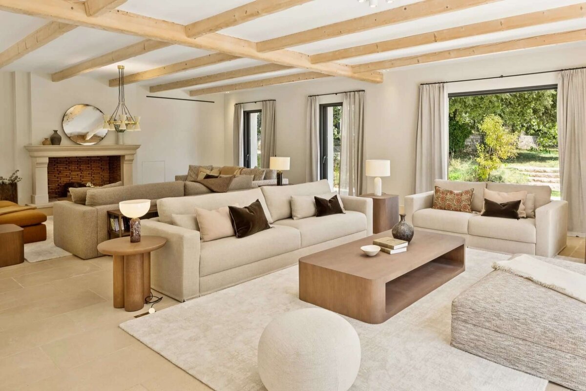 A spacious, modern living room with exposed wood beams, neutral sofas, a fireplace, and large windows looking out to lush greenery.