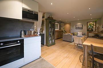 An open-plan living space features a modern kitchen on the left, blending into a living area with a sofa and a dining table, all on light wooden floors.