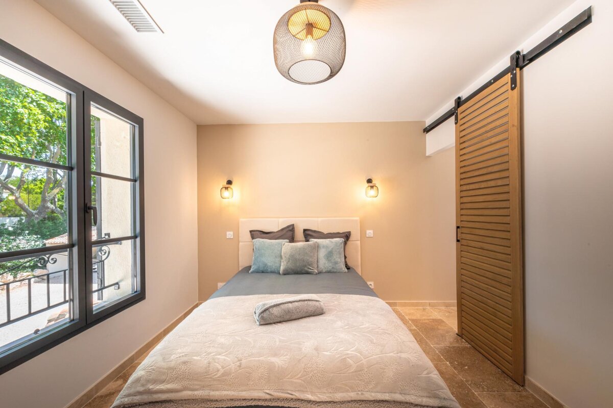 A modern, inviting bedroom with a bed, serene blue pillows, a large window showcasing trees, warm lighting, a unique sliding barn door, and light-colored walls.