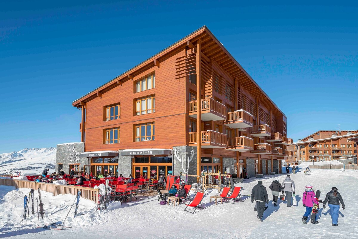 A modern wooden ski lodge with a bustling outdoor restaurant on a sunny, snowy day. People walk on a snow path with mountains in the distance.