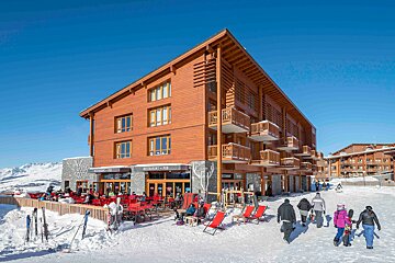 A modern wooden ski lodge with a bustling outdoor restaurant on a sunny, snowy day. People walk on a snow path with mountains in the distance.