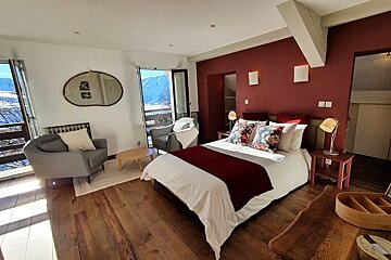 A cozy bedroom features a red accent wall, dark wood floors, a large bed, and a seating area. A balcony overlooks scenic snowy mountains.
