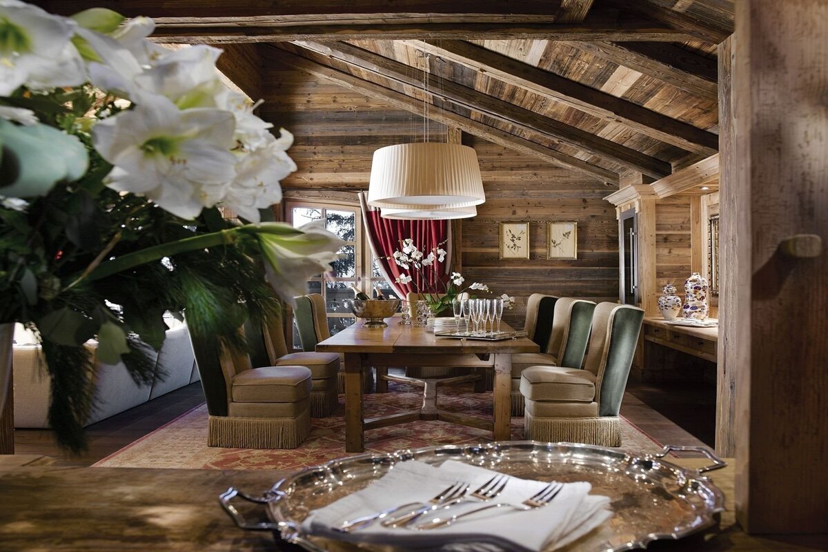 A rustic wooden chalet dining room features a long table set with champagne flutes and comfy chairs. White flowers and a silver tray are in the foreground.